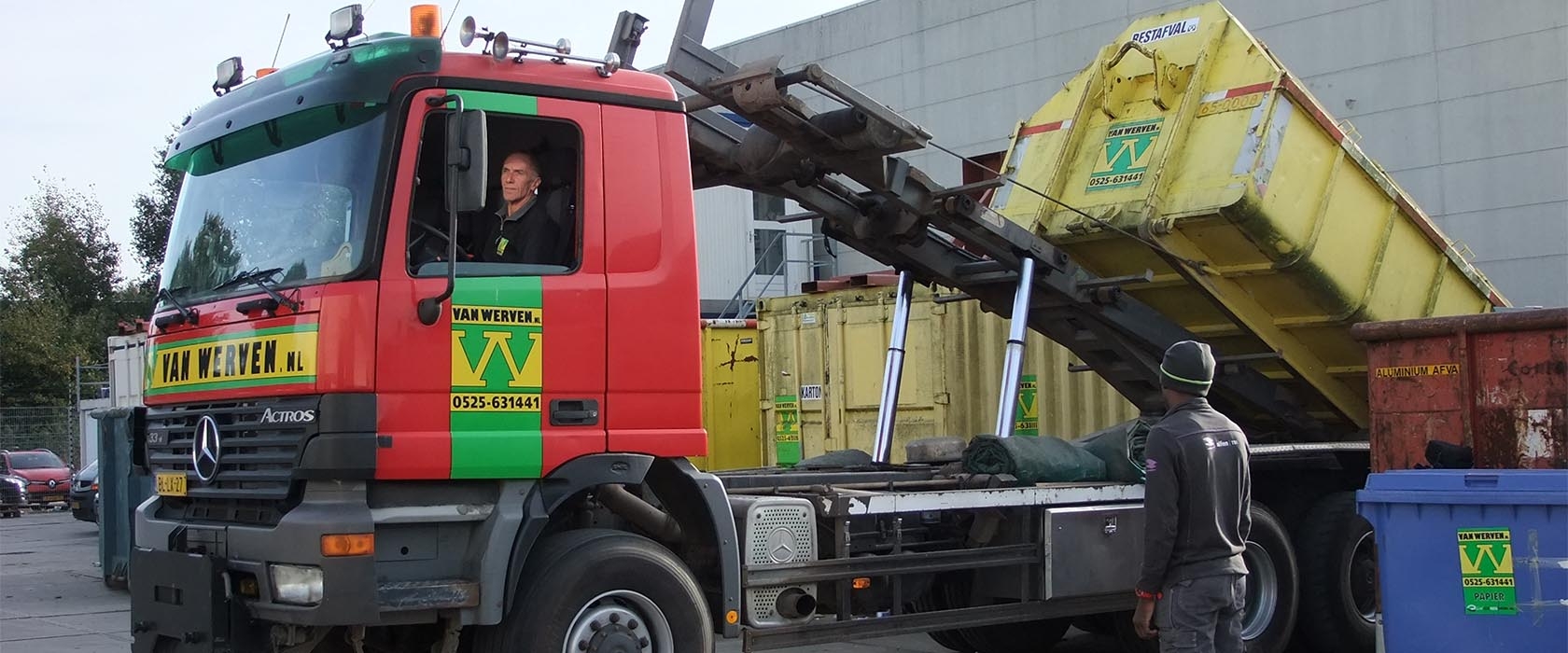 Een containerauto van Van Werven kantelt een gele zeecontainer op het terrein van Alfen, naast containers voor karton, restafval en aluminium
