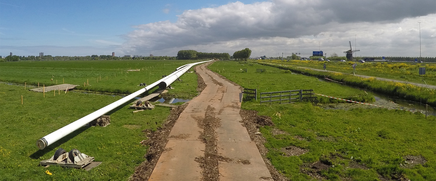 De tijdelijke rijbaan loopt door het weiland naast de witte gasleiding die verlegd wordt, met op de achtergrond de snelweg en een molen