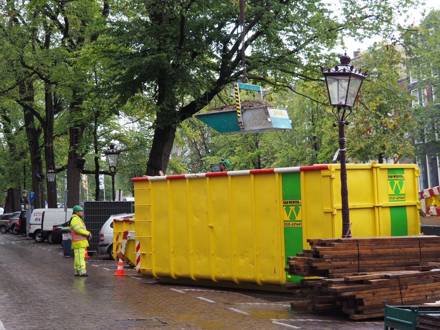 Een kleine puinbak hangt aan een kraan boven een grote gele Van Werven-container aan de gracht, terwijl een verkeersregelaar in geel hesje toekijkt